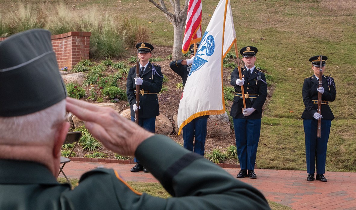 A veteran salutes during the Veterans Memorial Garden dedication ceremony as NC State’s color guard presents the colors, with the memorial’s landscaped grounds and brick architectural elements visible in the background.